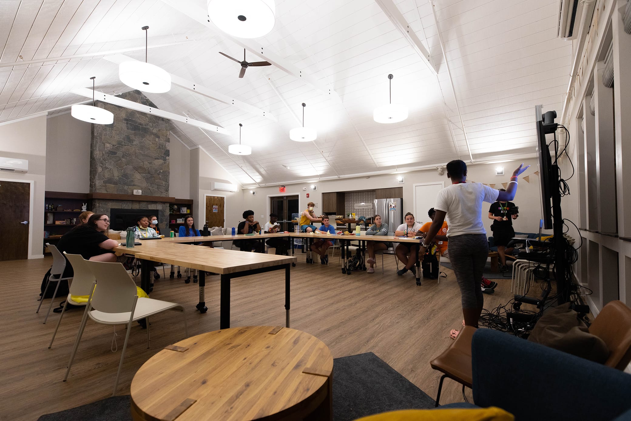 A group of people sits around a large U-shaped table in a modern, well-lit meeting room, while one person stands and presents in front of them.