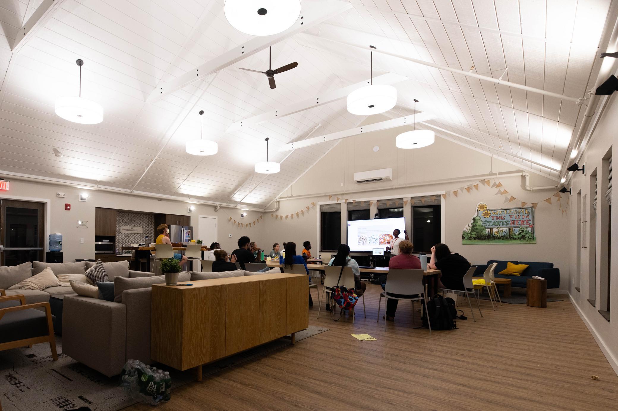 A group of people sit in a brightly lit room watching a presentation on a screen. The room has modern furniture, high ceilings, and decorative lights.