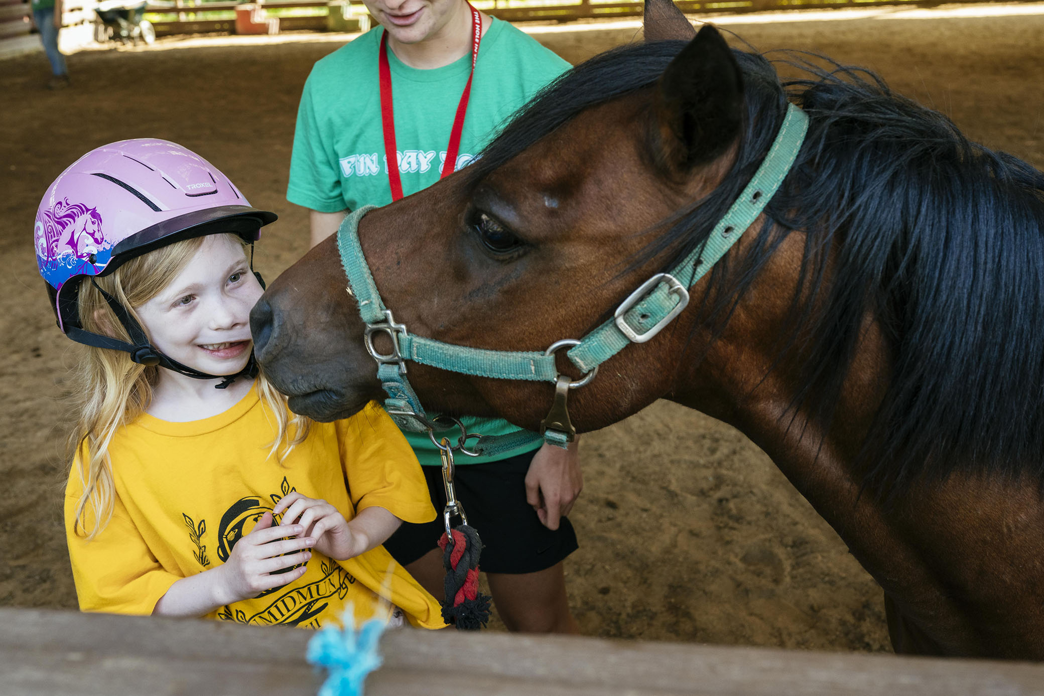 A smiling girl in a yellow shirt and pink helmet stands next to a brown horse, which is nuzzling her cheek. Another person in a green shirt stands beside them, holding the horses reins.