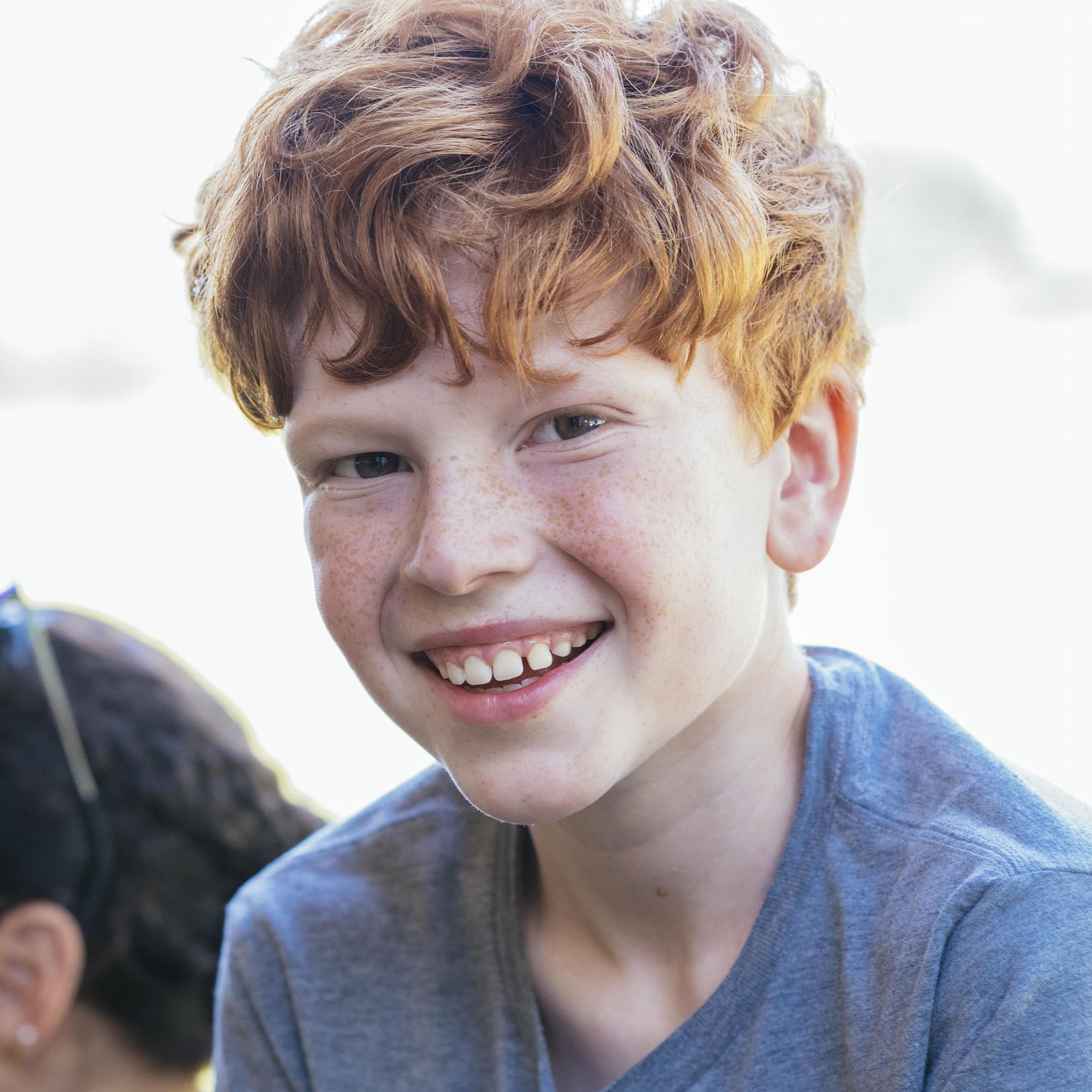 A young boy with curly red hair and freckles smiles at the camera, wearing a gray shirt. The background is bright and slightly blurred.