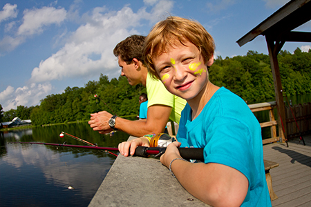 A young boy with face paint smiles at the camera while holding a fishing rod on a dock. An older boy fishes beside him. Trees and a lake are in the background. Its a bright, sunny day.