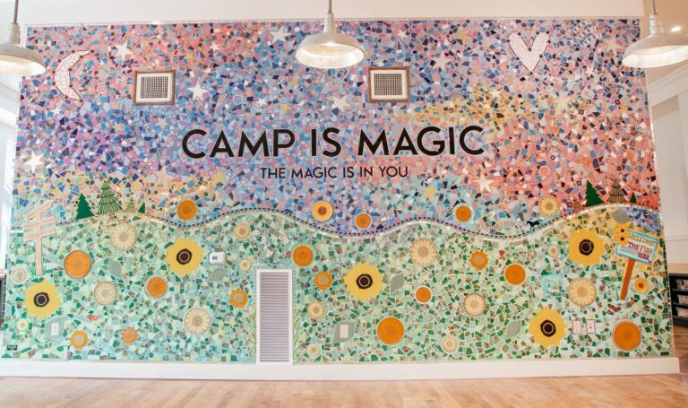 Colorful mosaic mural with text CAMP IS MAGIC, THE MAGIC IS IN YOU over a landscape with flowers, trees, stars, and patterns. Three hanging lights and a wood floor are visible.