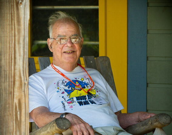 An older man with glasses sits smiling on a wooden chair outside. He wears a colorful T-shirt, shorts, and a red lanyard. The background features a yellow door and green siding.