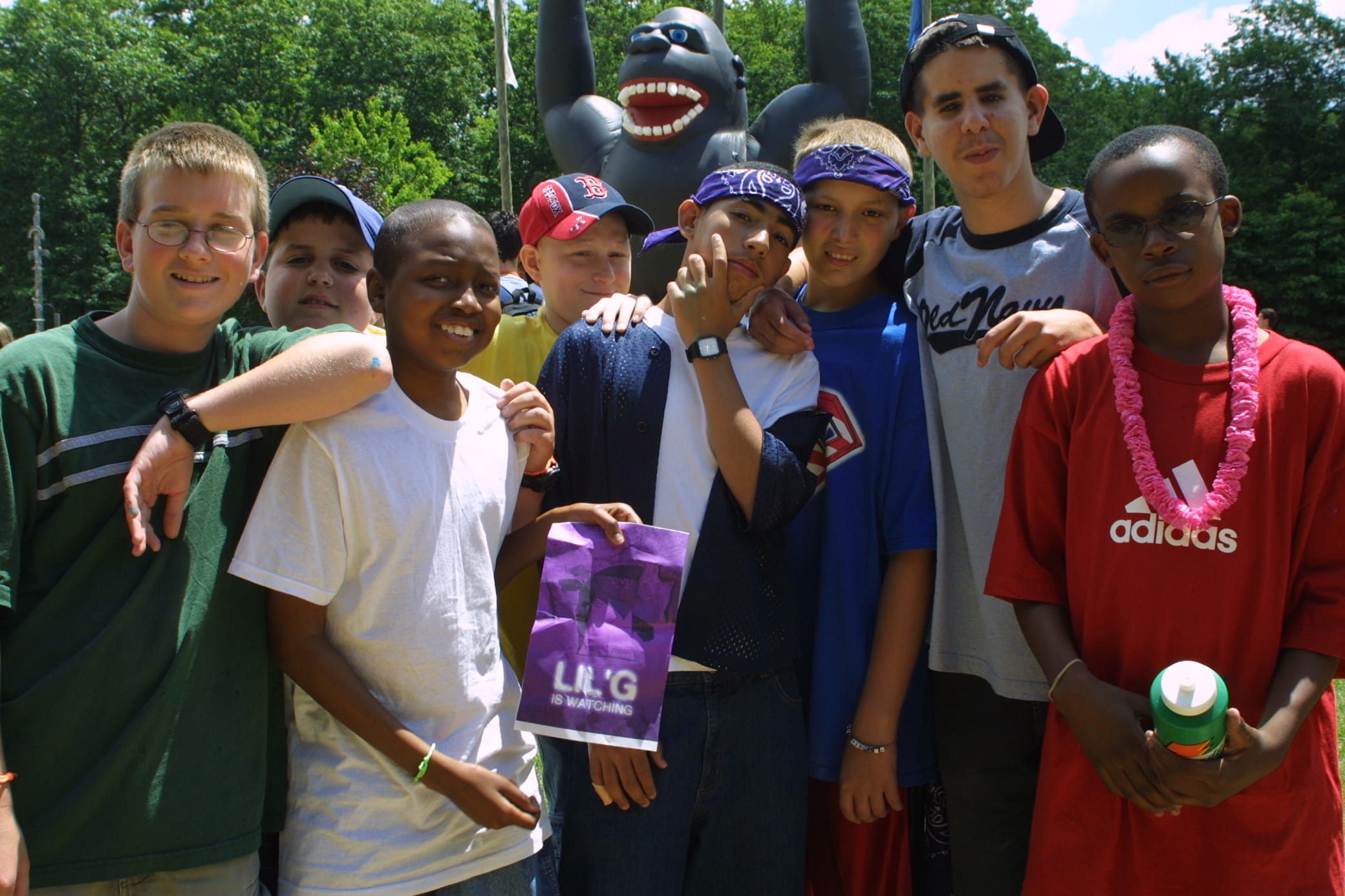 A group of boys pose together outdoors, some smiling and some making playful gestures, with a large inflatable gorilla and trees in the background.