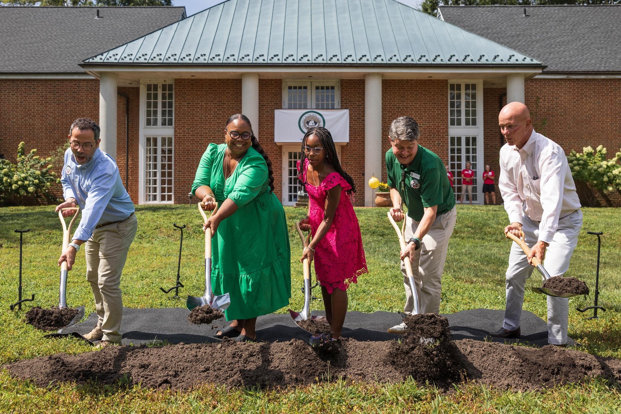 Five people in colorful attire smile while holding shovels and digging into soil at a groundbreaking ceremony on a lawn, with a brick building in the background.