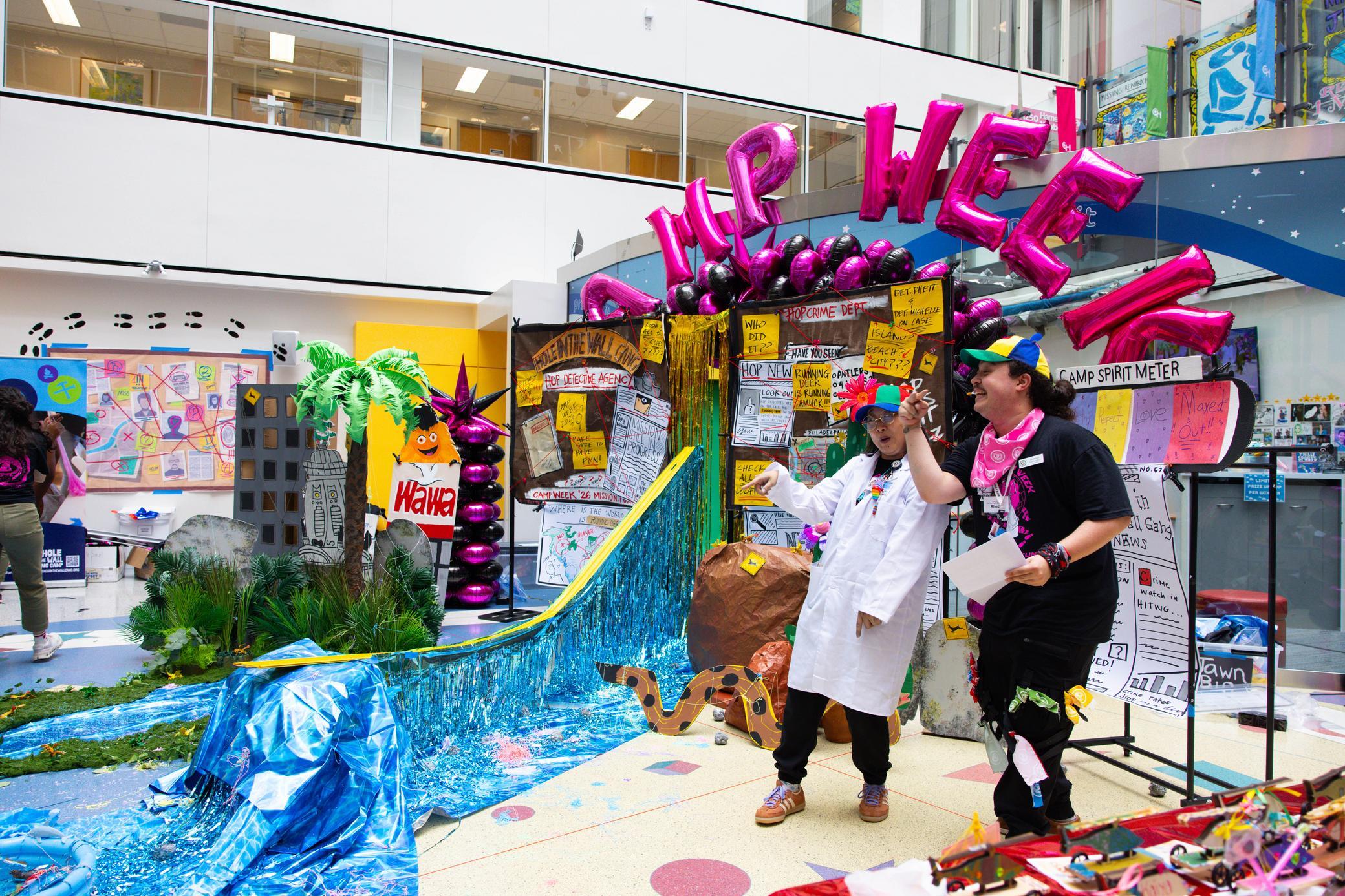 Two people in costumes stand in a colorful, decorated indoor space with CAMP WEEK balloons, posters, a blue waterfall, fake plants, and props, creating a lively, festive atmosphere.