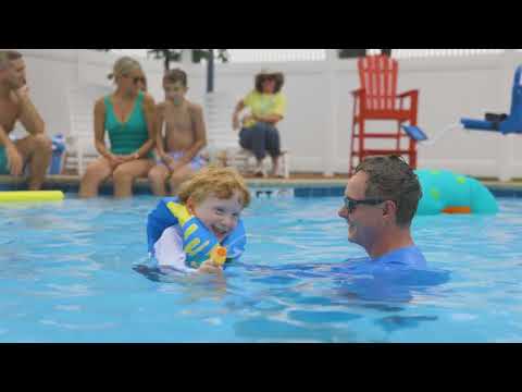 A young child wearing a life jacket smiles while playing in a pool with an adult. Other people sit nearby watching. The scene is bright and cheerful.
