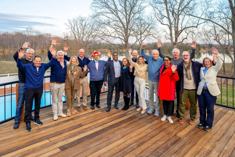 A group of fifteen diverse adults standing on a wooden deck outdoors, all smiling and waving with their hands raised.