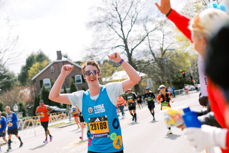 A smiling marathon runner in sunglasses raises his arms in celebration while passing cheering supporters; other runners and a house are visible in the background on a sunny day.